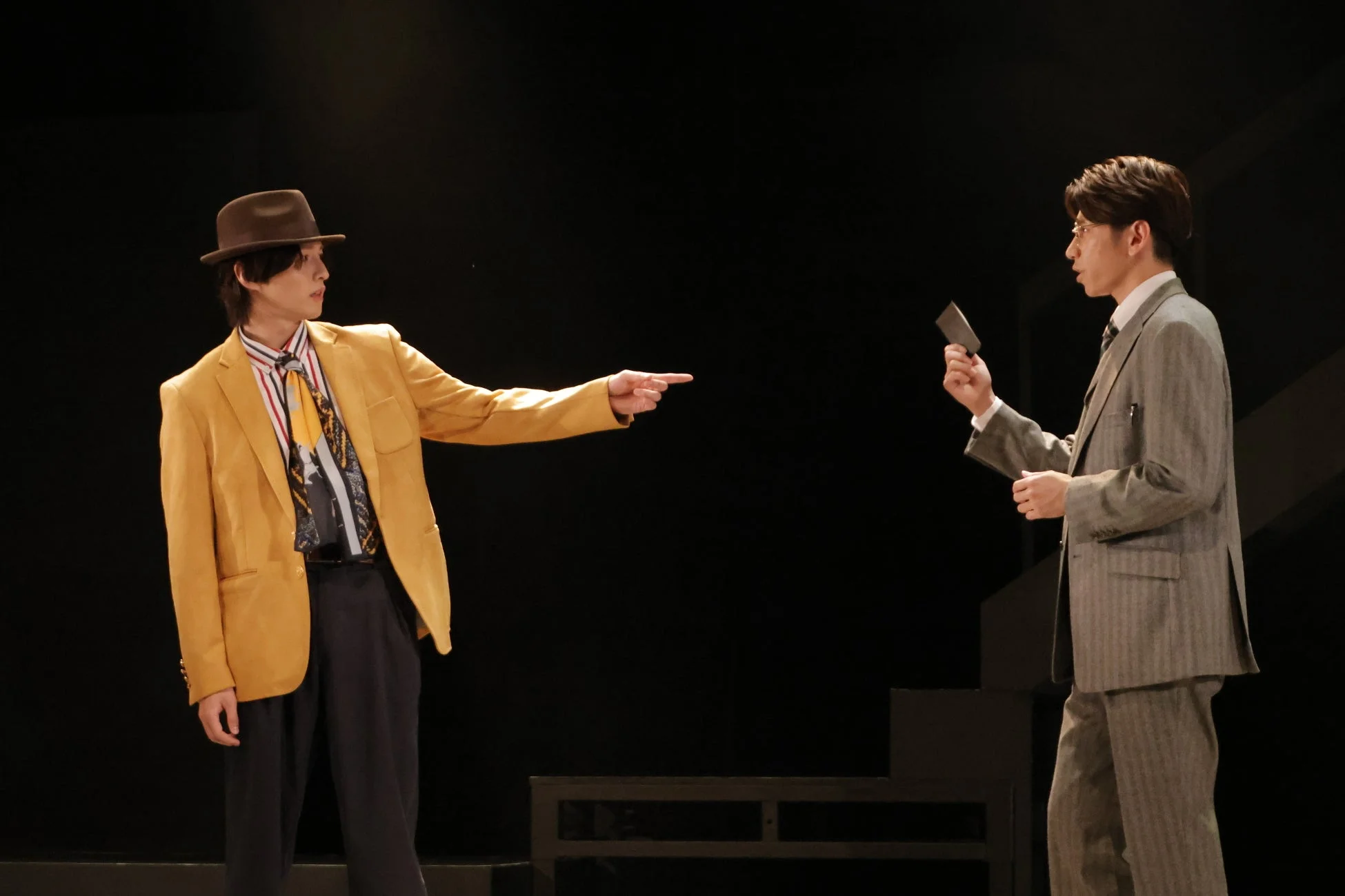 暗い舞台上で、黄色のジャケットと帽子をかぶった男性が、グレーのスーツを着た男性に指を指している