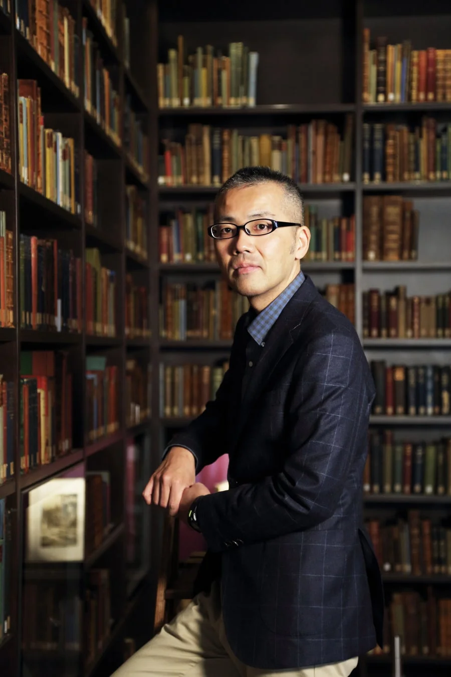 本棚が並ぶ図書館で、メガネをかけた男性がカメラの方を向いて立っている