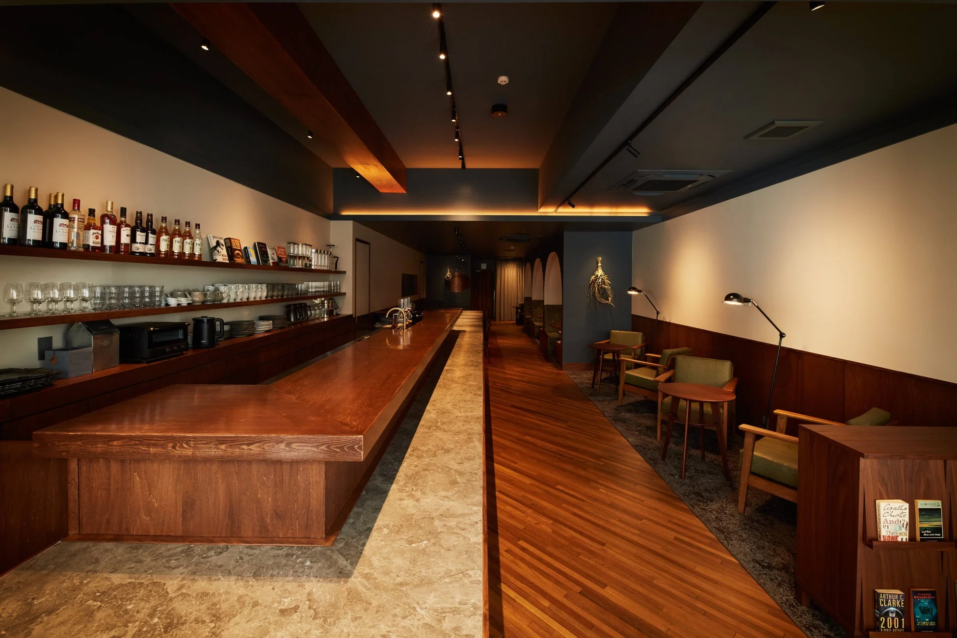 ボトルやグラスが並ぶ長い木製カウンターと、ゆったりと座れるアームチェアが配置された、モダンで落ち着いた雰囲気のバーまたはラウンジの店内