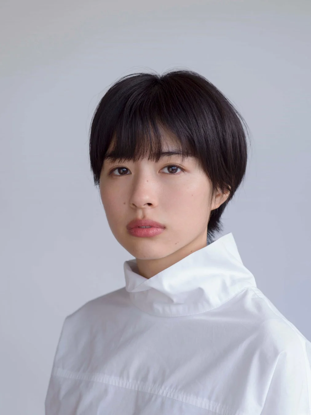 白いハイネックシャツを着た若い女性のポートレートです。黒いショートヘアで、カメラをまっすぐ見つめており、表情は穏やかです。背景は無地の明るいグレーです。