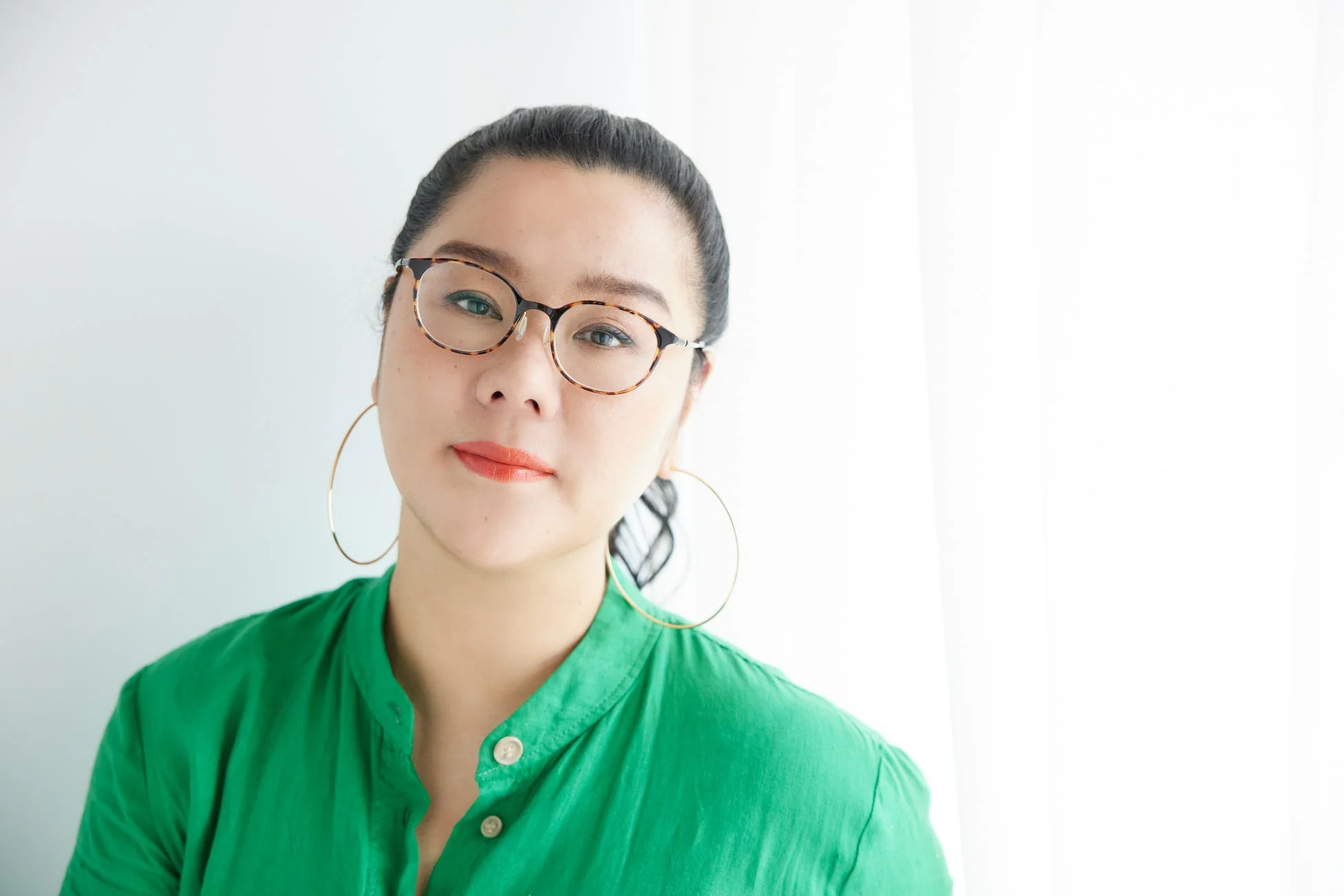 白い背景を背に、べっ甲柄の眼鏡と緑色のシャツを身につけた女性のポートレートです。髪をまとめ、大きなフープピアスをつけ、カメラをまっすぐに見つめています。
