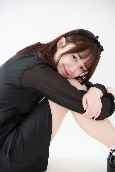 白い背景の前で、黒いワンピースを着た若い女性が座り込み、膝を抱えるようにして優しく微笑んでいるポートレート写真