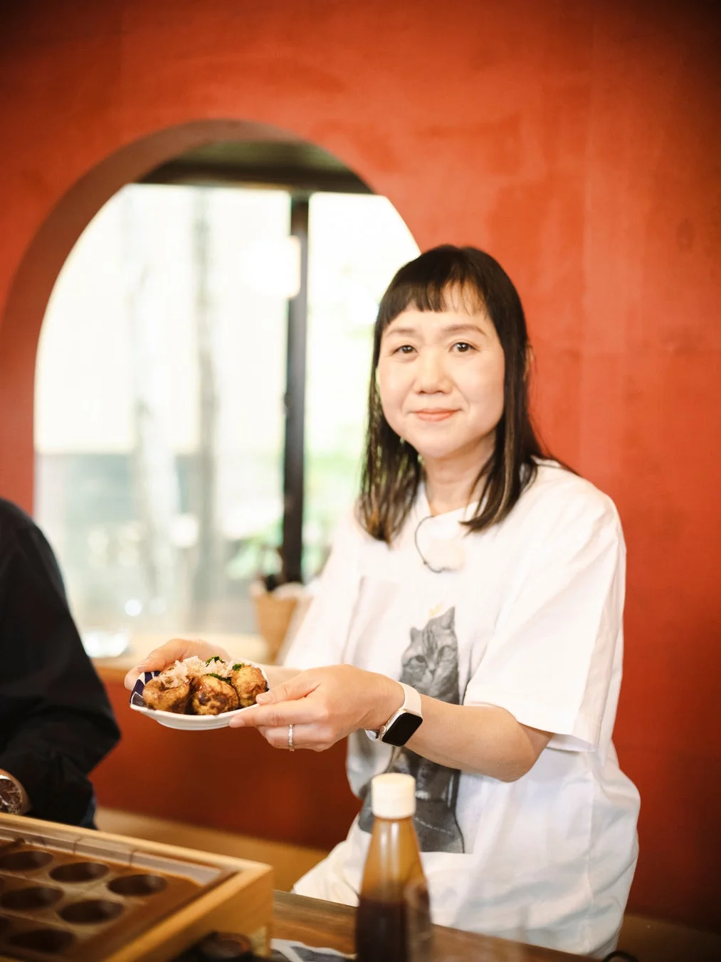笑顔の女性が、たこ焼きが乗った皿を両手で差し出している。背景は赤い壁で、窓から光が差し込む明るい室内で、和やかな食事が楽しまれている様子が伺える。