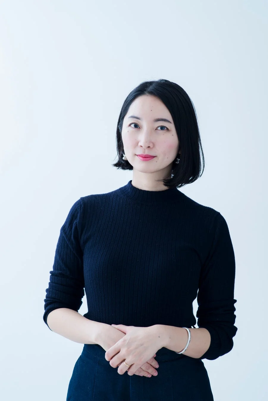 白い背景の前で、黒いリブニットを着た黒髪ショートヘアの女性がカメラを見つめているポートレート写真です。両手を組んでおり、落ち着いた印象を与えます。
