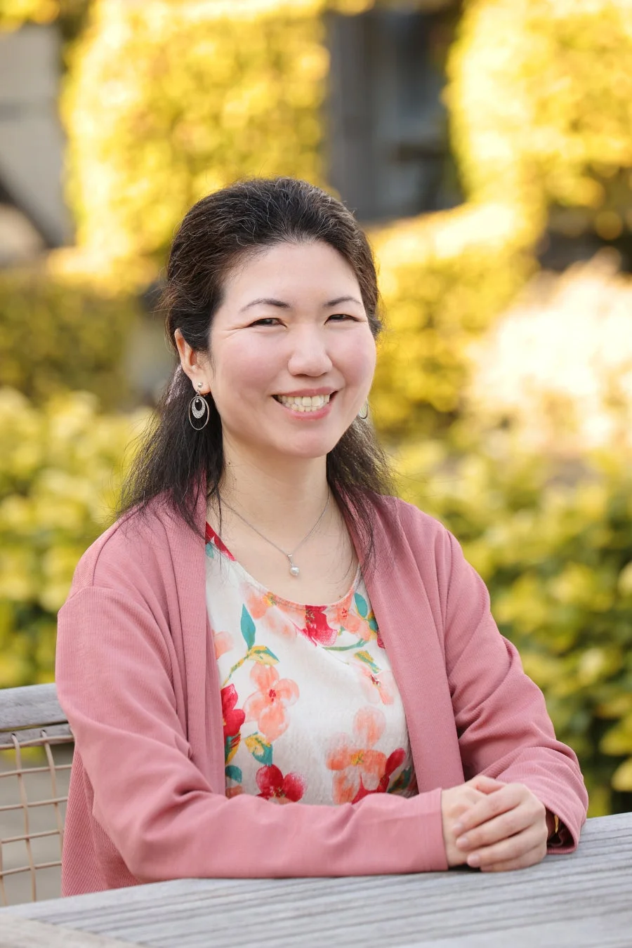 屋外で笑顔を見せるアジア人女性のポートレート。ピンクのカーディガンと花柄のトップスを着用し、木製のテーブルに手を組んで座っています。背景はぼかされた黄色い植物で、明るく親しみやすい雰囲気の画像です。